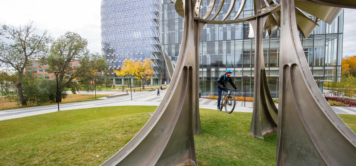 A close up shot through a statue on UCalgary campus shows a cyclist and a building in the distance