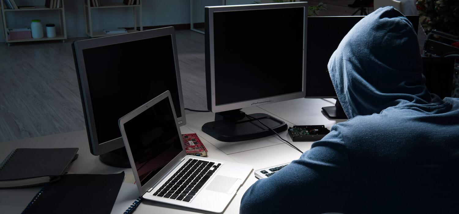 A hooded figure sits in front of multiple computers